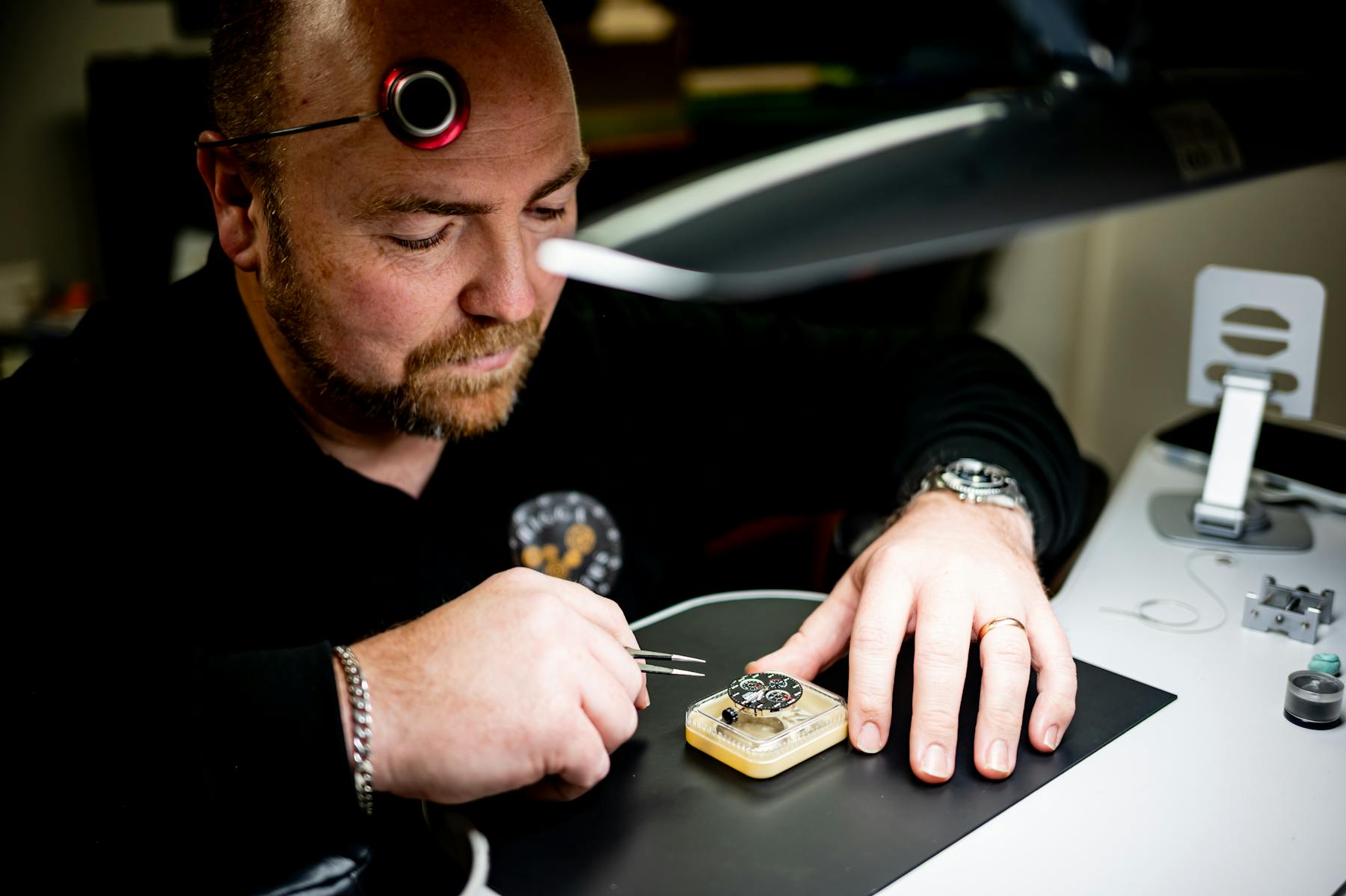 A watchmaker works on a precision mechanical watch at a workshop bench.